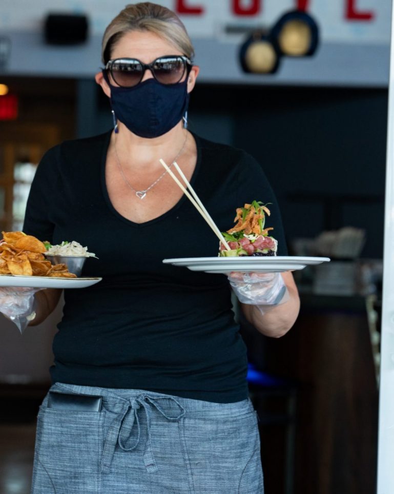 A masked waitress carrying two plates of food to a table