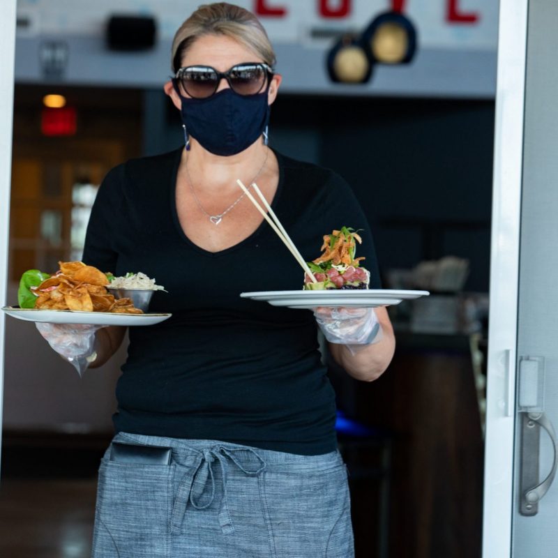 A masked waitress carrying two plates of food to a table